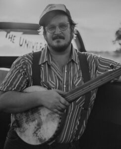 man with hat on holding a banjo poses for a photo behind a truck