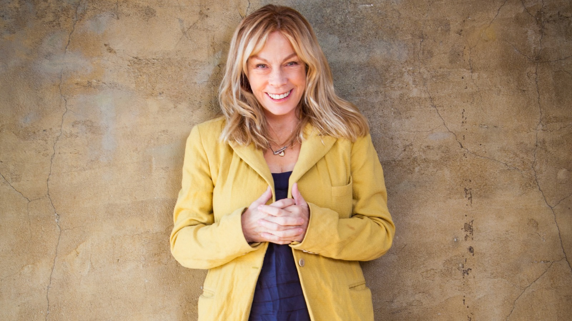 woman in yellow jacket poses in front of a natural toned wall