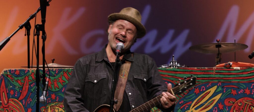 man wearing hat and playing guitar sings into mic