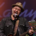 man wearing hat and playing guitar sings into mic